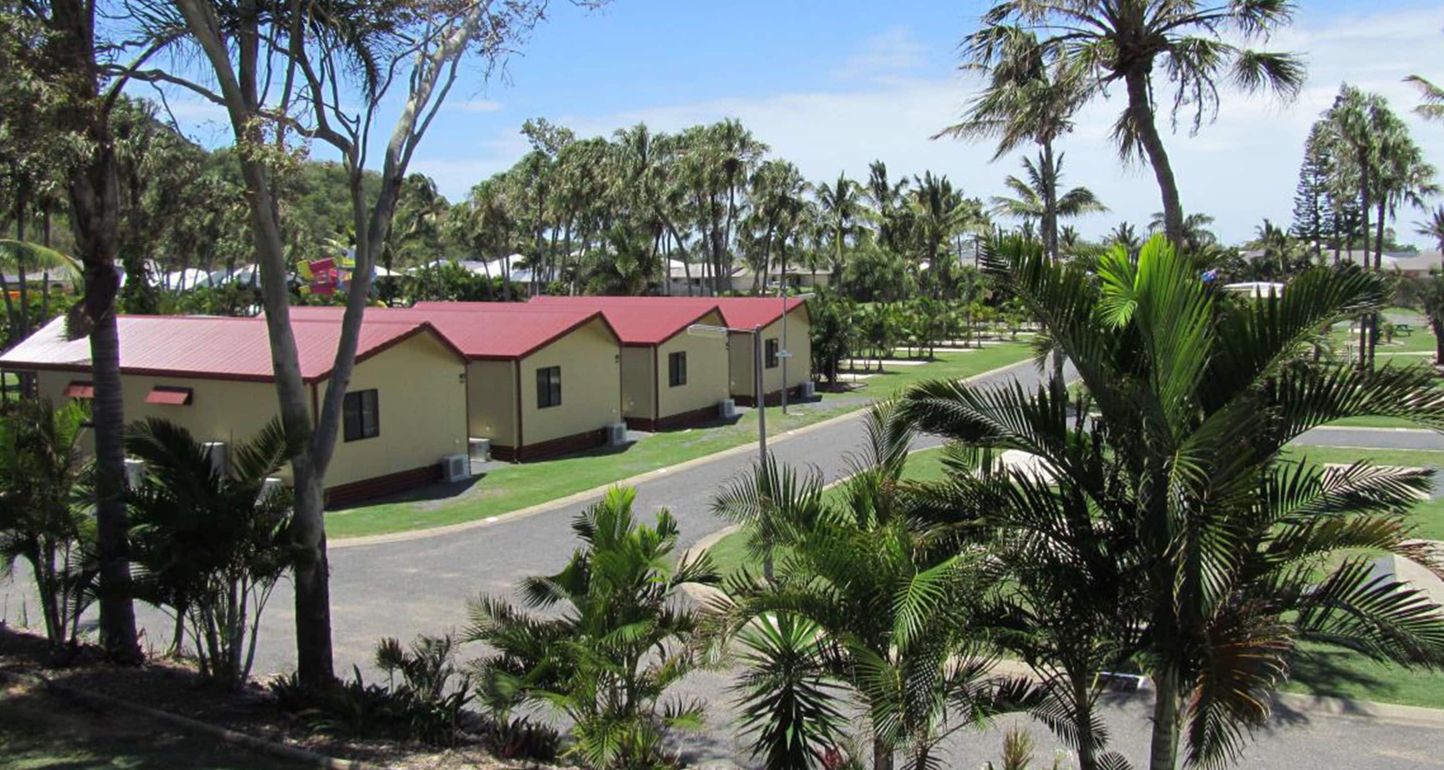 A row of single-story buildings with red roofs, surrounded by palm trees and greenery.