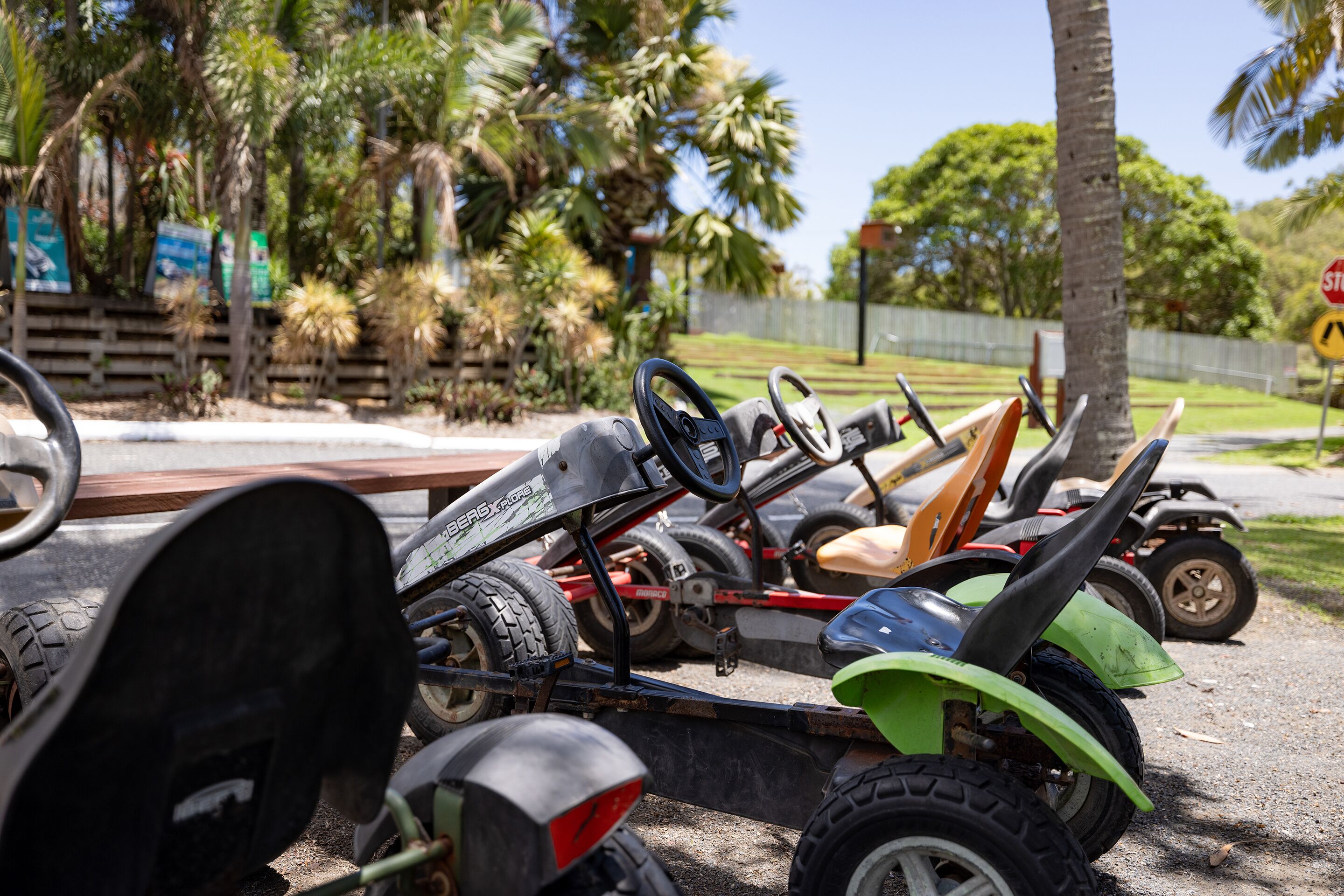 Una fila de karts de carrera estacionados en un área pavimentada, con palmeras y una cerca de madera al fondo.