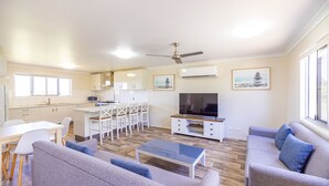 Una sala de estar moderna con sofá, mesa de centro y televisor. Hay una zona de comedor con sillas y una mesa, y una cocina con armarios y estufa.