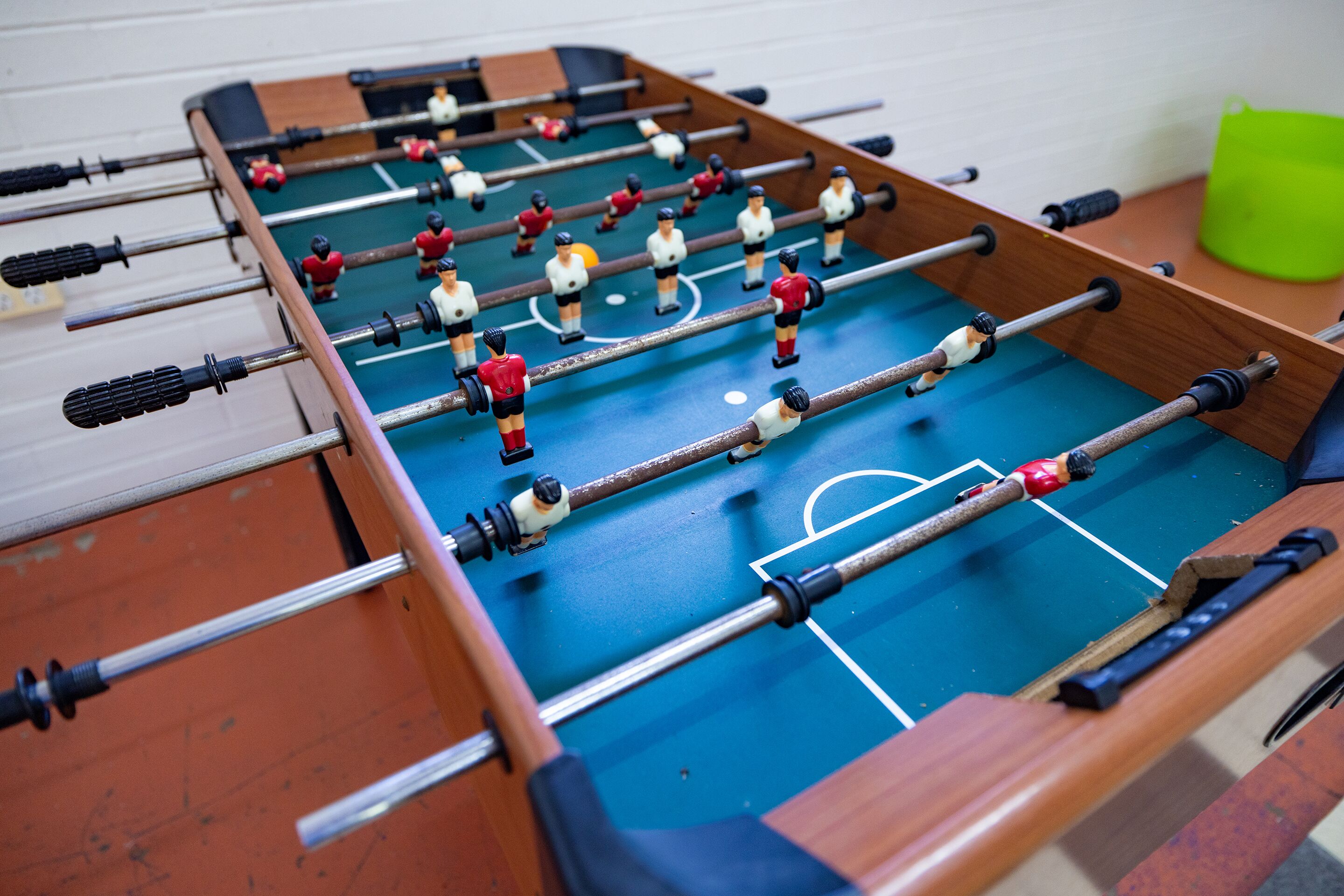 A foosball table with players in red and white uniforms, a green bucket, and a wooden table with a blue playing surface.