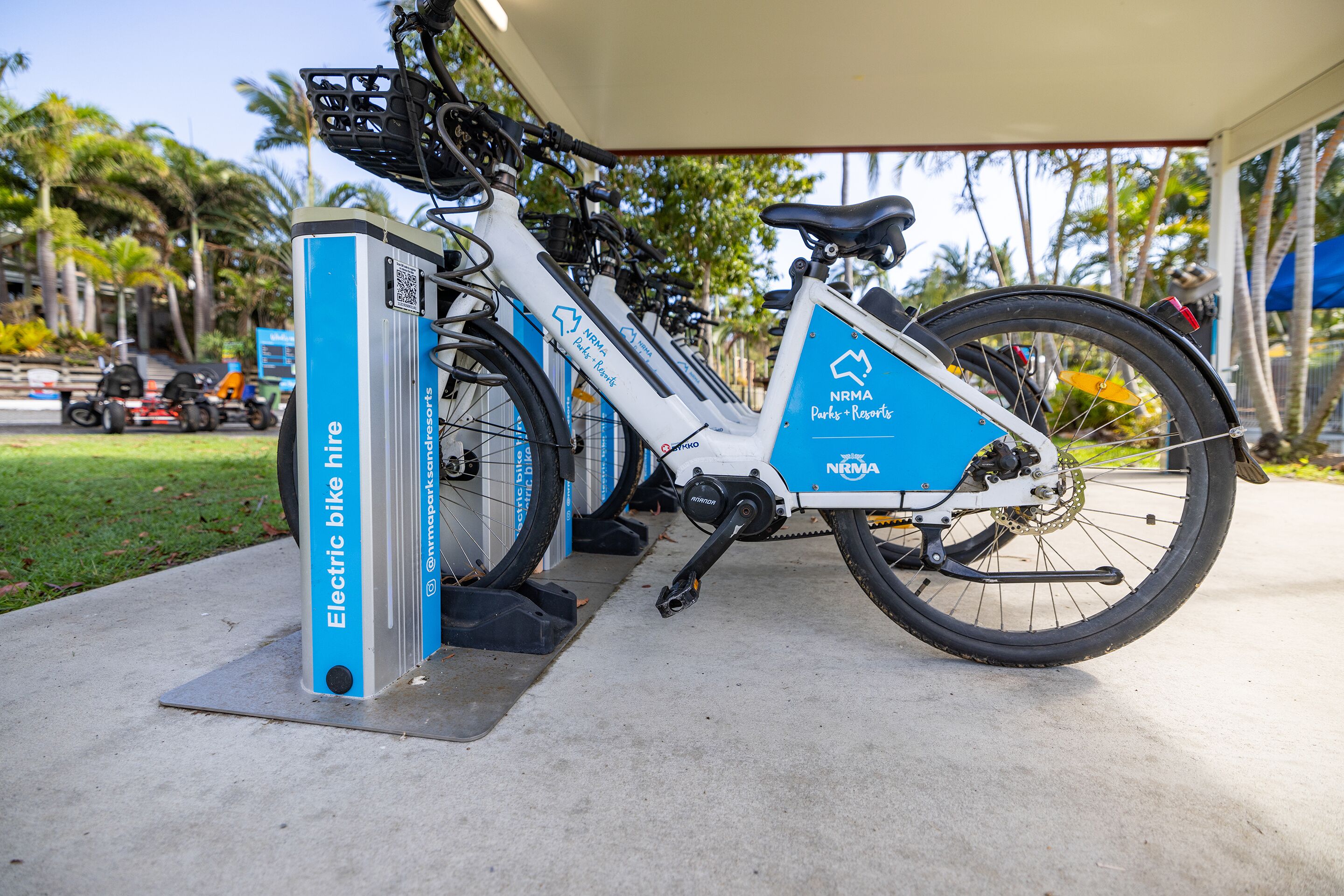 Elektrische fietsen geparkeerd bij een station, met een helder uitzicht op de omgeving, inclusief een weg en palmbomen.