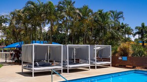 Zona de piscina con cuatro camas con dosel blanco, una piscina azul cristalina y palmeras al fondo.