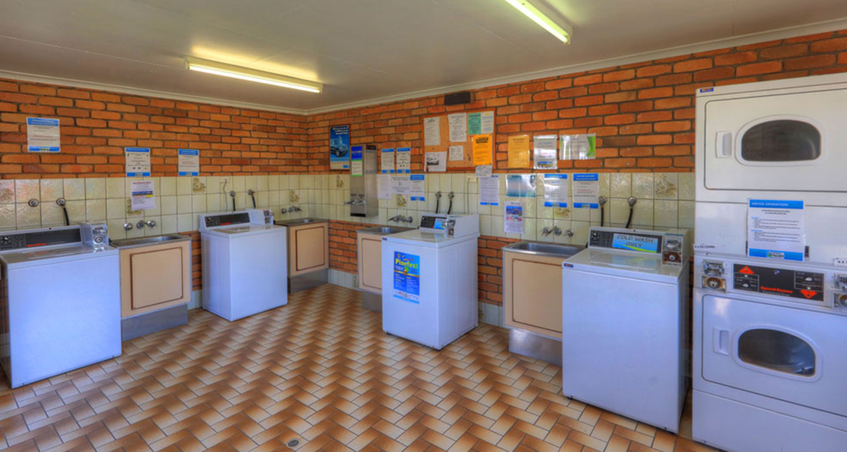 A laundry facility with multiple washing machines and dryers, tiled walls, and a brick floor.