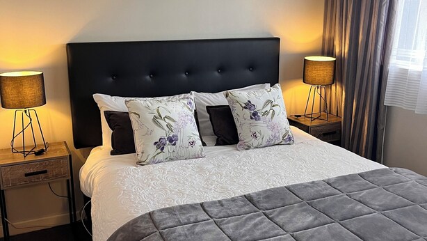 A neatly made bed with a dark headboard, two bedside lamps, and a window with curtains. - Aorangi Motel (Fairlie)