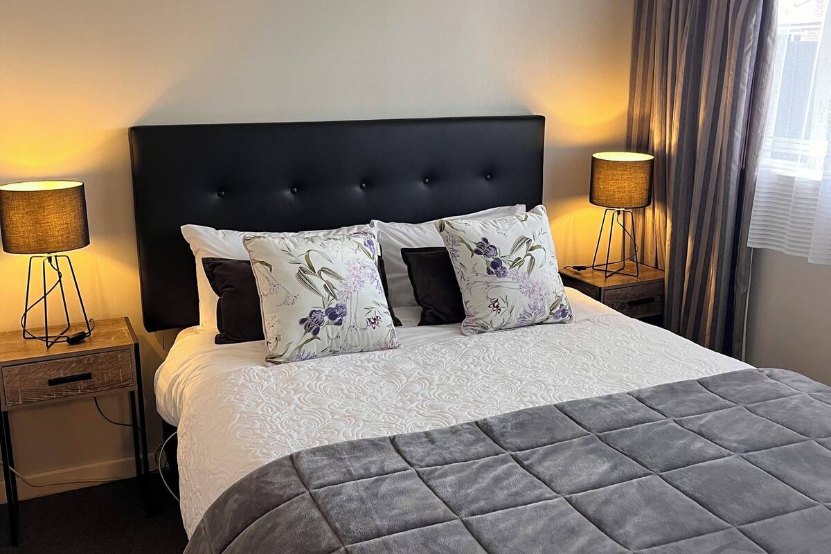 A neatly made bed with a dark headboard, two bedside lamps, and a window with curtains.