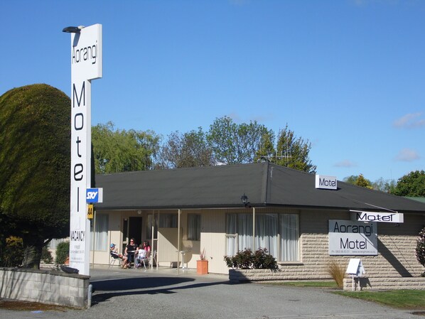 A motel with a signboard, a small outdoor seating area, and a building with a dark roof. - Aorangi Motel (Fairlie)