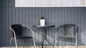 A patio with two metal chairs and a small table, a potted plant, and a window with white curtains. - Aorangi Motel (Fairlie)