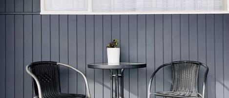 A patio with two metal chairs and a small table, a potted plant, and a window with white curtains.