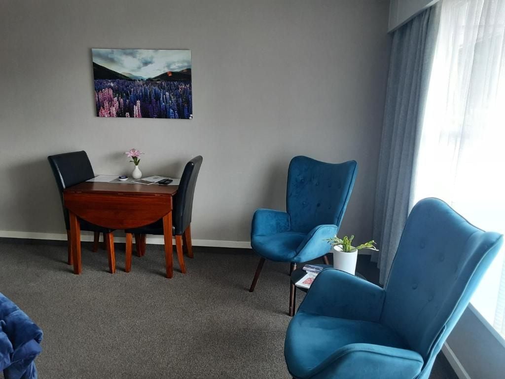A room with a dining table, two chairs, a blue armchair, a potted plant, and a window with curtains.