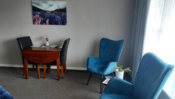 A room with a dining table, two chairs, a blue armchair, a potted plant, and a window with curtains.