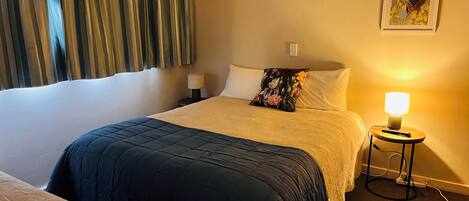 A hotel room with a large bed, a side table with a lamp, a wall-mounted light, and a framed picture on the wall.