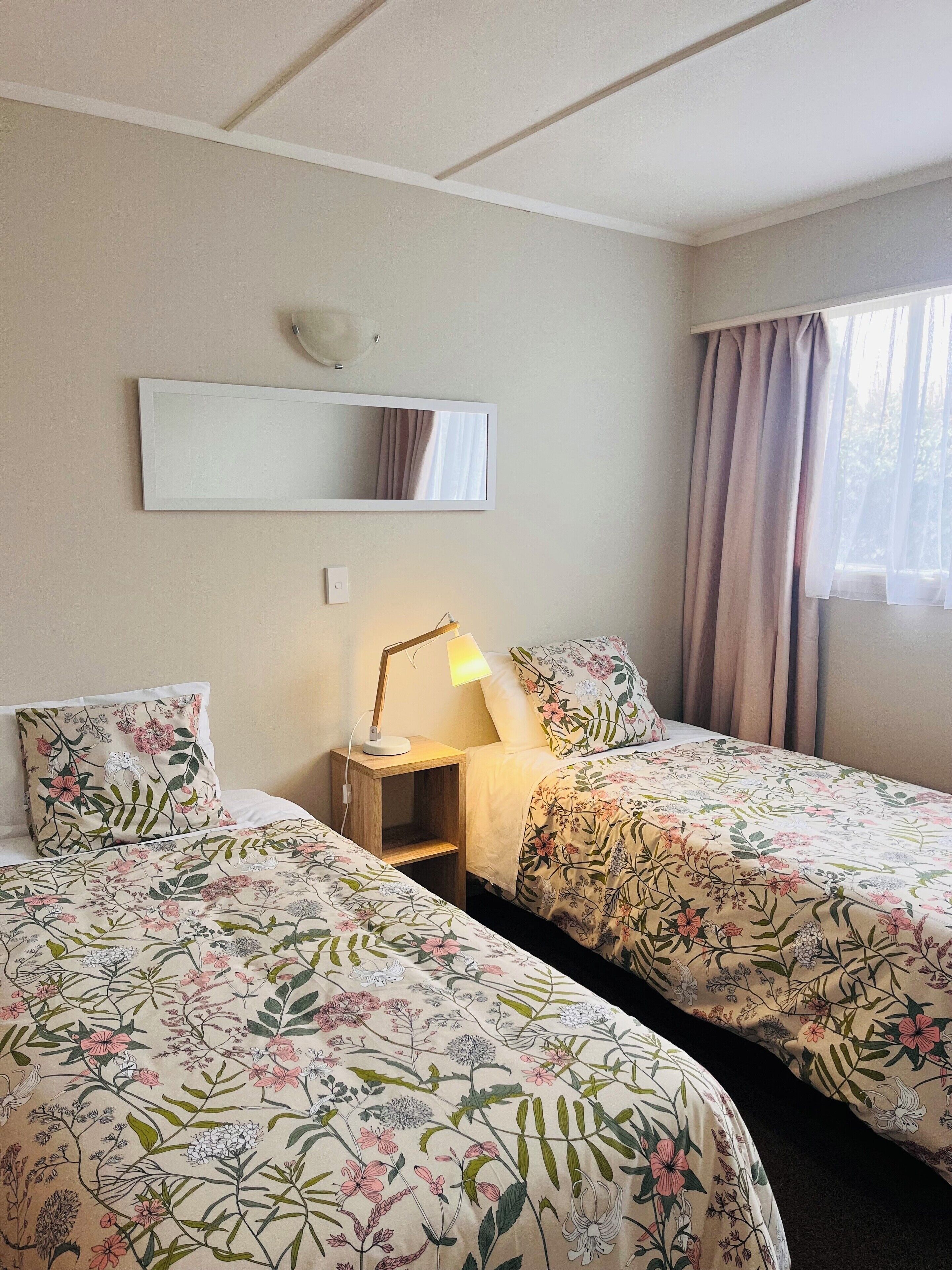 A hotel room with two beds, each with floral bedding, a bedside table with a lamp, a mirror on the wall, and a window with curtains.