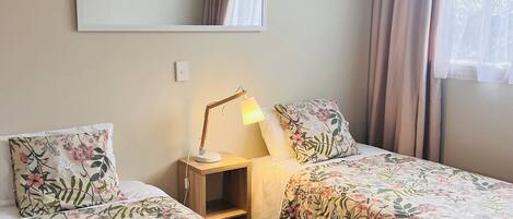 A hotel room with two beds, each with floral bedding, a bedside table with a lamp, a mirror on the wall, and a window with curtains.