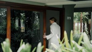 Une personne vêtue d’une longue robe blanche se tient devant une porte-fenêtre coulissante, d’où l’on aperçoit des plantes tropicales.
