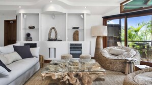 Un salon moderne avec une table basse en verre, un canapé, un fauteuil en osier et une grande fenêtre donnant sur un paysage verdoyant.