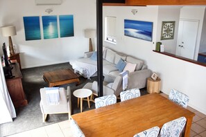 A living room with a wooden coffee table, a sofa, and a dining table with chairs. There are three paintings on the wall and a cabinet with a lamp.