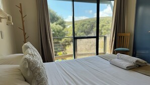 A neatly made bed with white linens, a wooden chair, and a view of greenery through large windows. - The Break, Margaret River Beach Houses (Gnarabup)