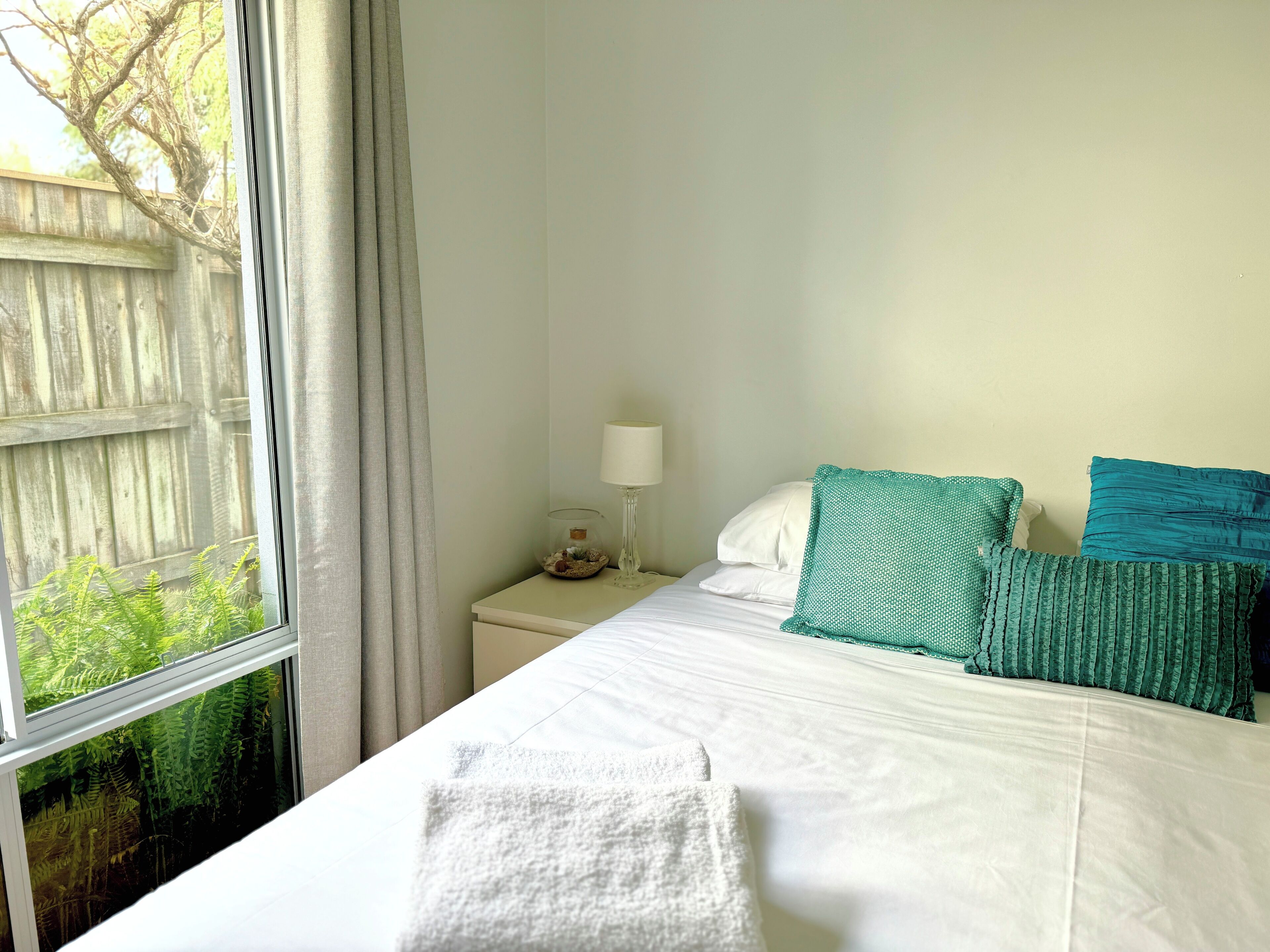 A neatly made bed with white linens and teal pillows, a bedside table with a lamp, and a window with a view of a wooden fence and greenery.