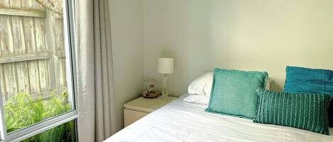A neatly made bed with white linens and teal pillows, a bedside table with a lamp, and a window with a view of a wooden fence and greenery.