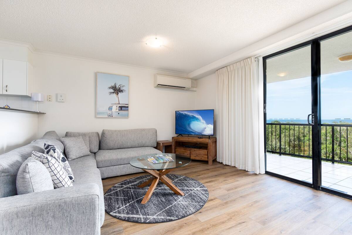 A modern living room with a grey sofa, a glass coffee table, a flat-screen TV, and a balcony with a view.