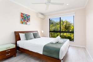 A neatly made bed with a green and white bedspread, a wooden headboard, and a nightstand with drawers. A colorful artwork is on the wall, and a large window offers a view of greenery outside. - Marcoola Beach Resort (Marcoola)