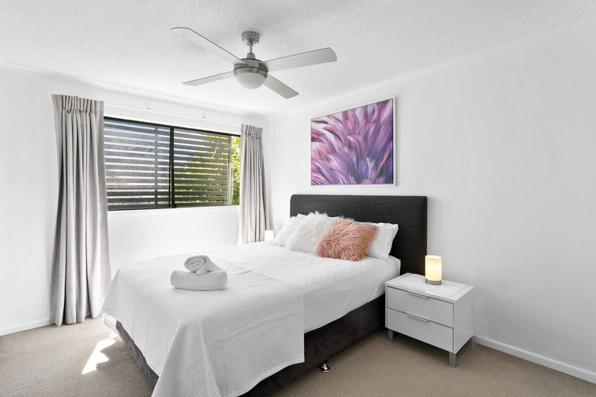 A bedroom with a bed, a nightstand, a ceiling fan, a window with blinds, and a wall-mounted artwork.