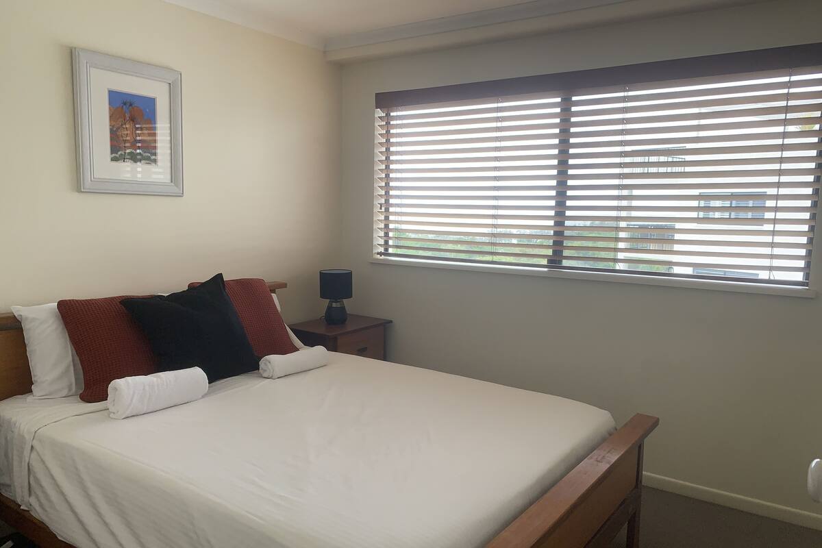 A neatly made bed with white linens and multiple pillows, a bedside table with a lamp, and a window with blinds.