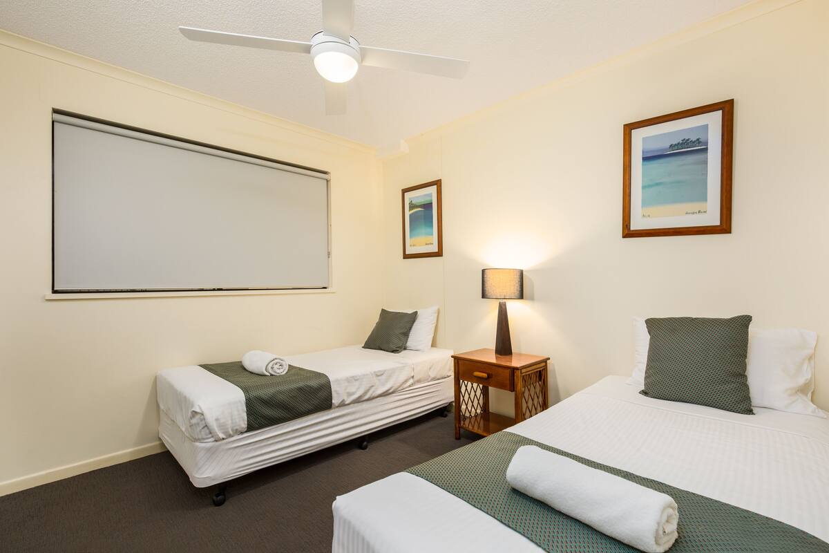 A hotel room with two beds, a ceiling fan, a wall-mounted TV, and a framed picture of a beach.