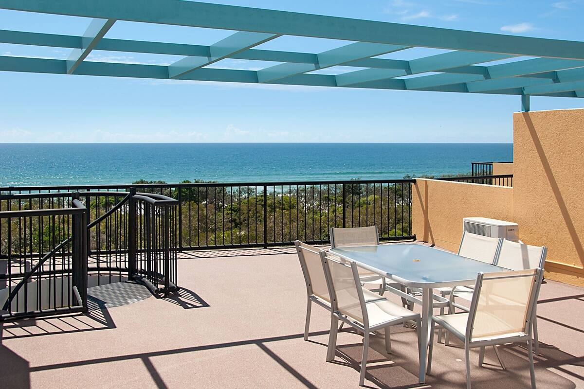 A balcony with a table and chairs, overlooking the ocean.