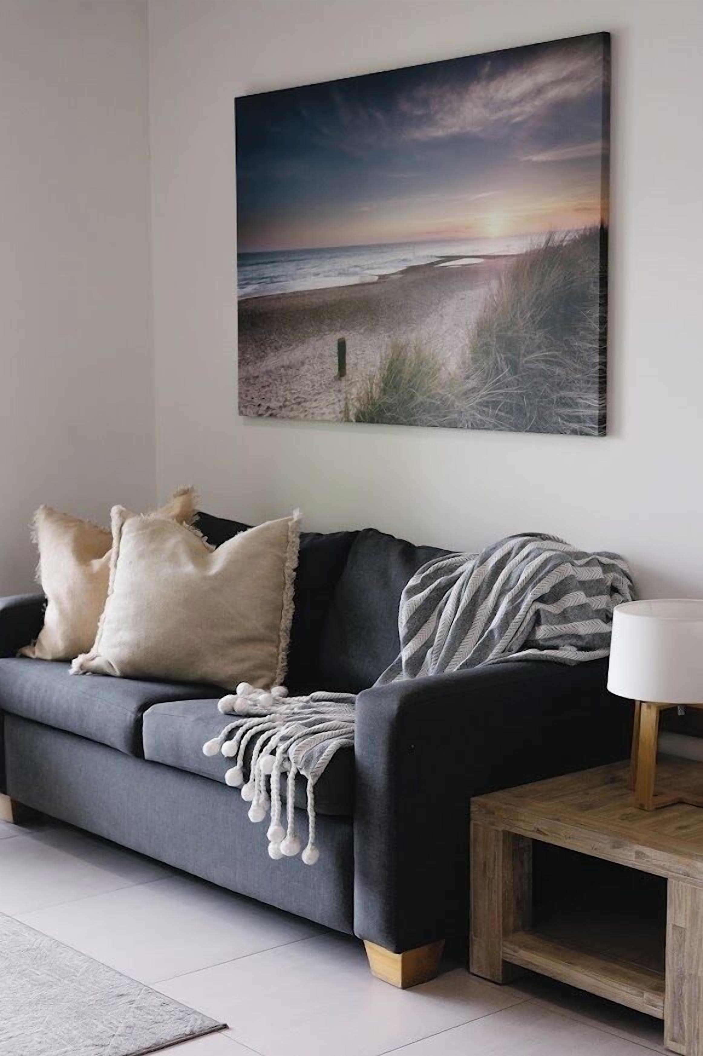 Ett vardagsrum med en soffa, kuddar, en filt, en lampa och en tavla som föreställer en strandvy.