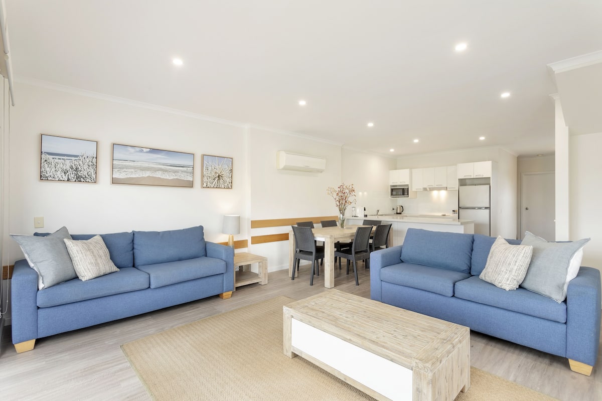 Uma sala de estar moderna com dois sofás azuis, uma mesa de centro de madeira e uma área de refeições com uma mesa e cadeiras de madeira.