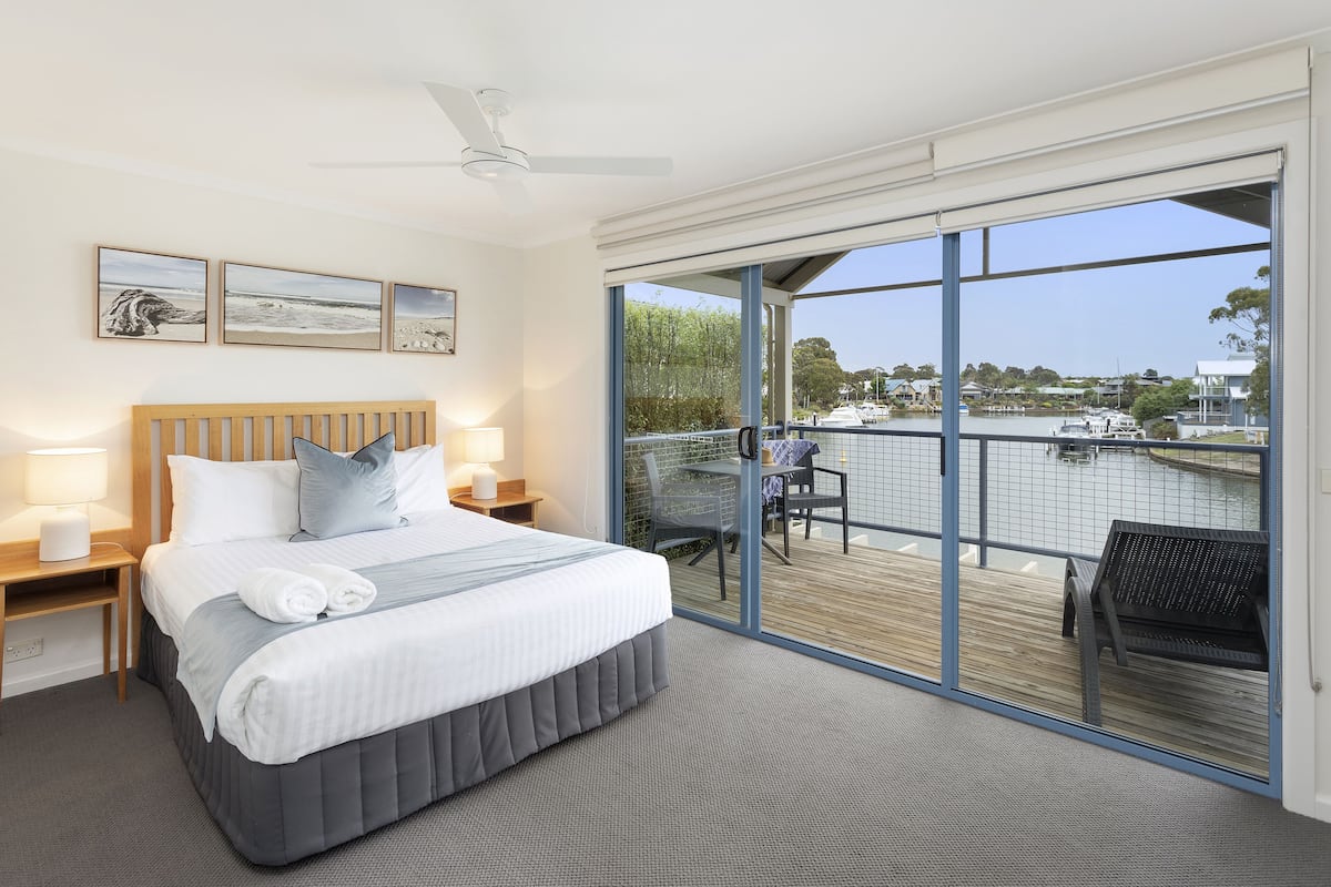 Quarto de hotel com uma cama grande, varanda com vista para a água e área de refeições com mesas ao ar livre.