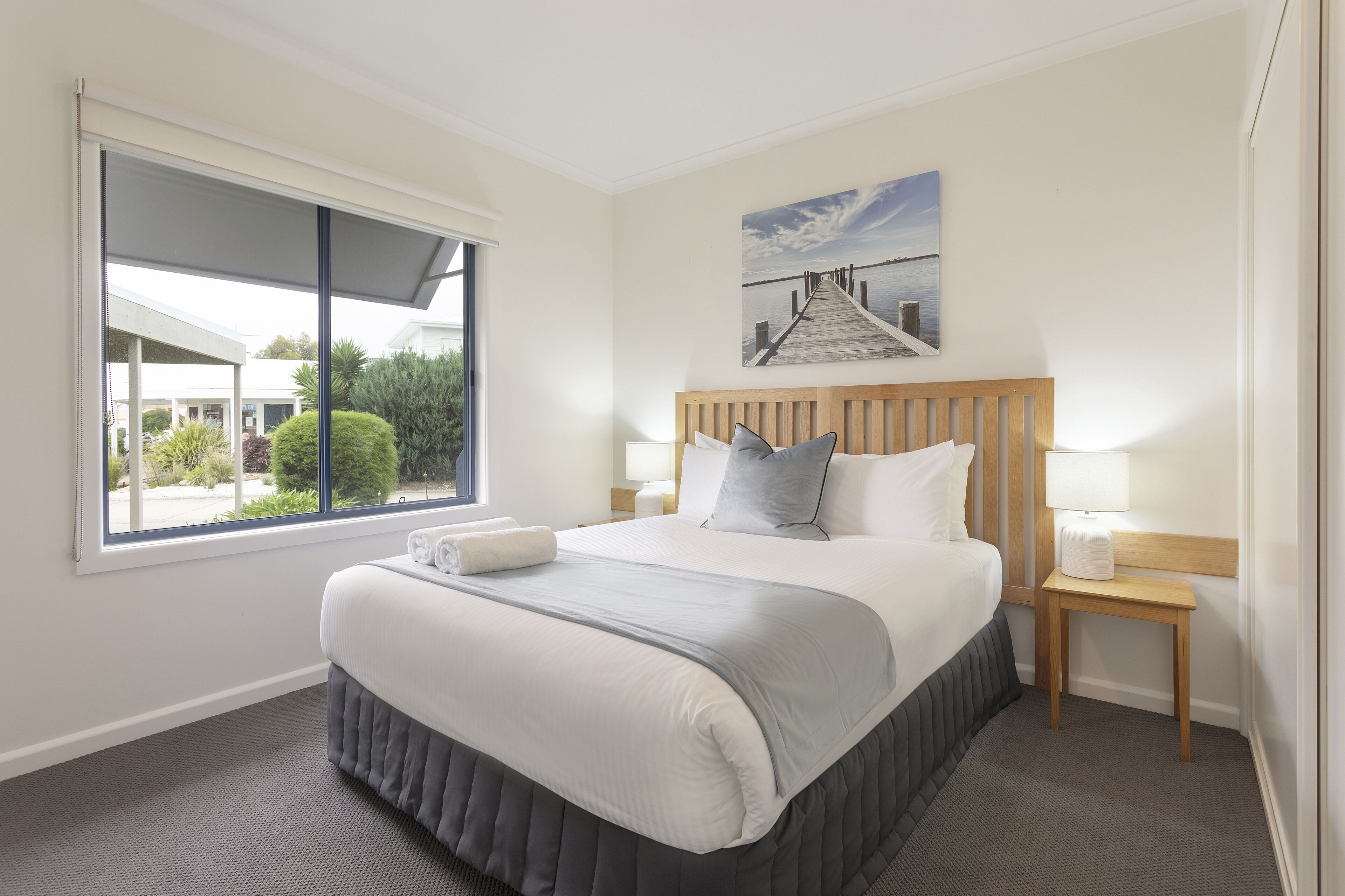 A neatly made bed with a wooden headboard, two bedside lamps, and a large window with a view of greenery and buildings outside.
