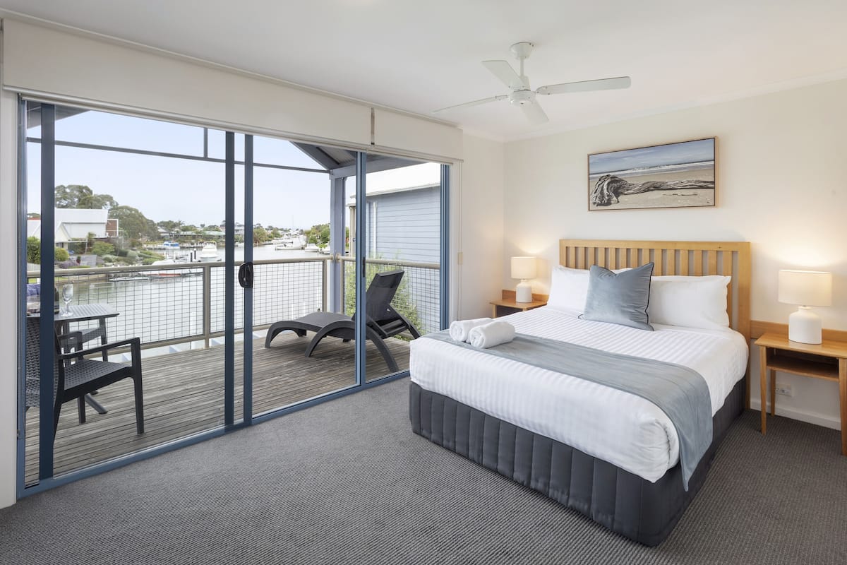 Quarto de hotel com uma grande porta de vidro deslizante que dá para uma varanda, uma cama com cabeceira estofada cinza e um quadro na parede.