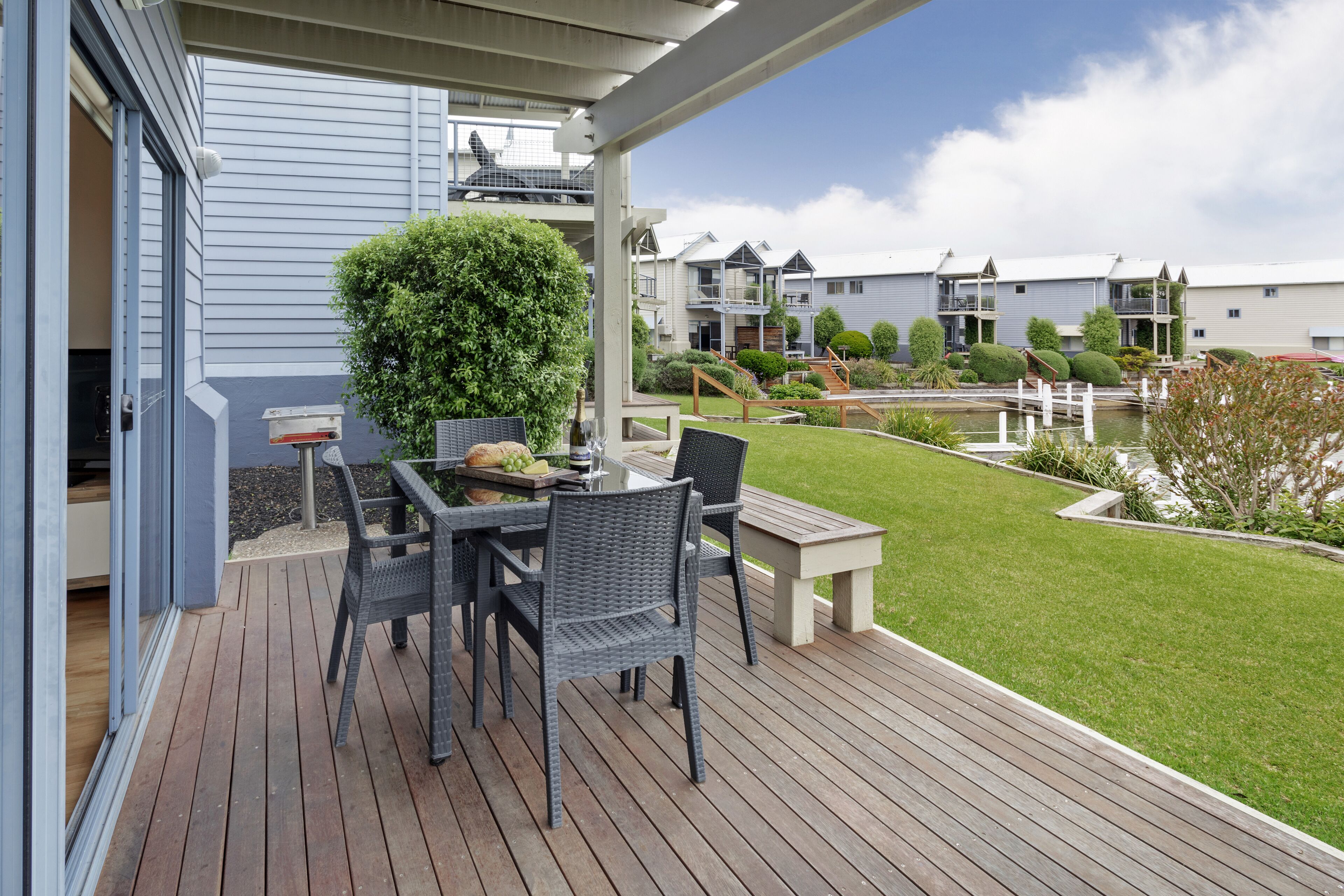 Una terraza de madera con mesa y sillas, un pequeño jardín y vistas a edificios residenciales.