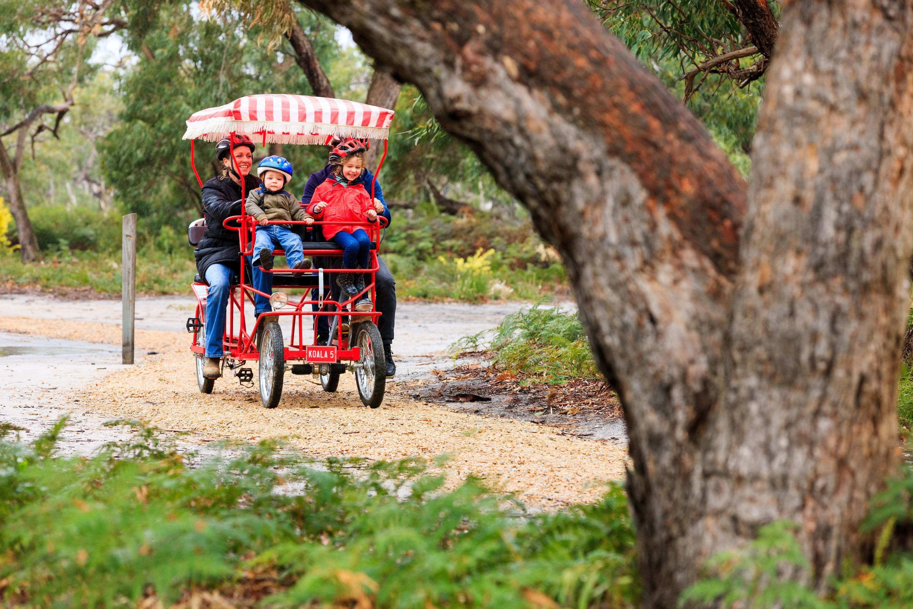 A red and white covered cart with three passengers, including a child, traveling on a dirt path surrounded by trees.