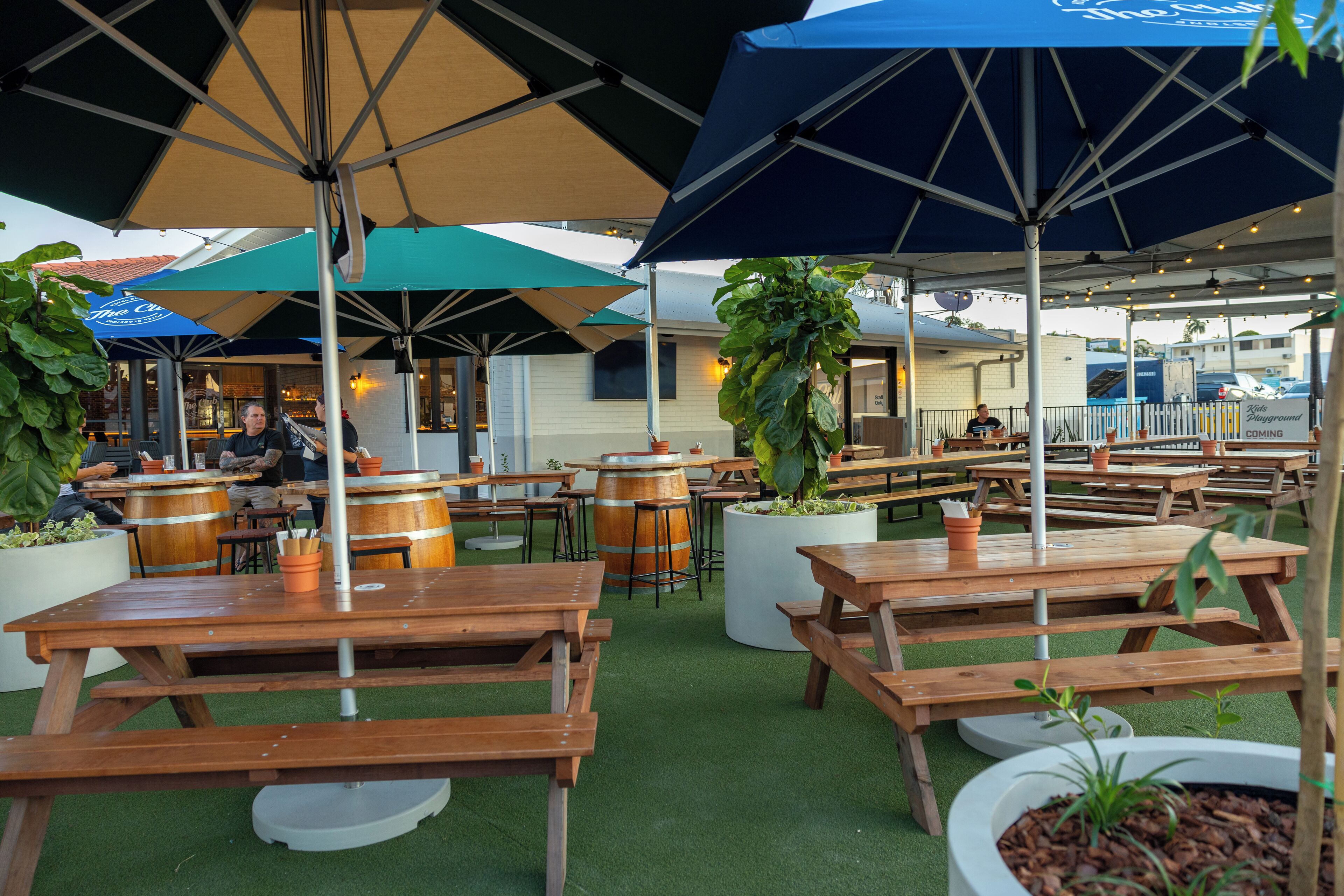 An outdoor patio area with wooden picnic tables and umbrellas, surrounded by potted plants and barrels.