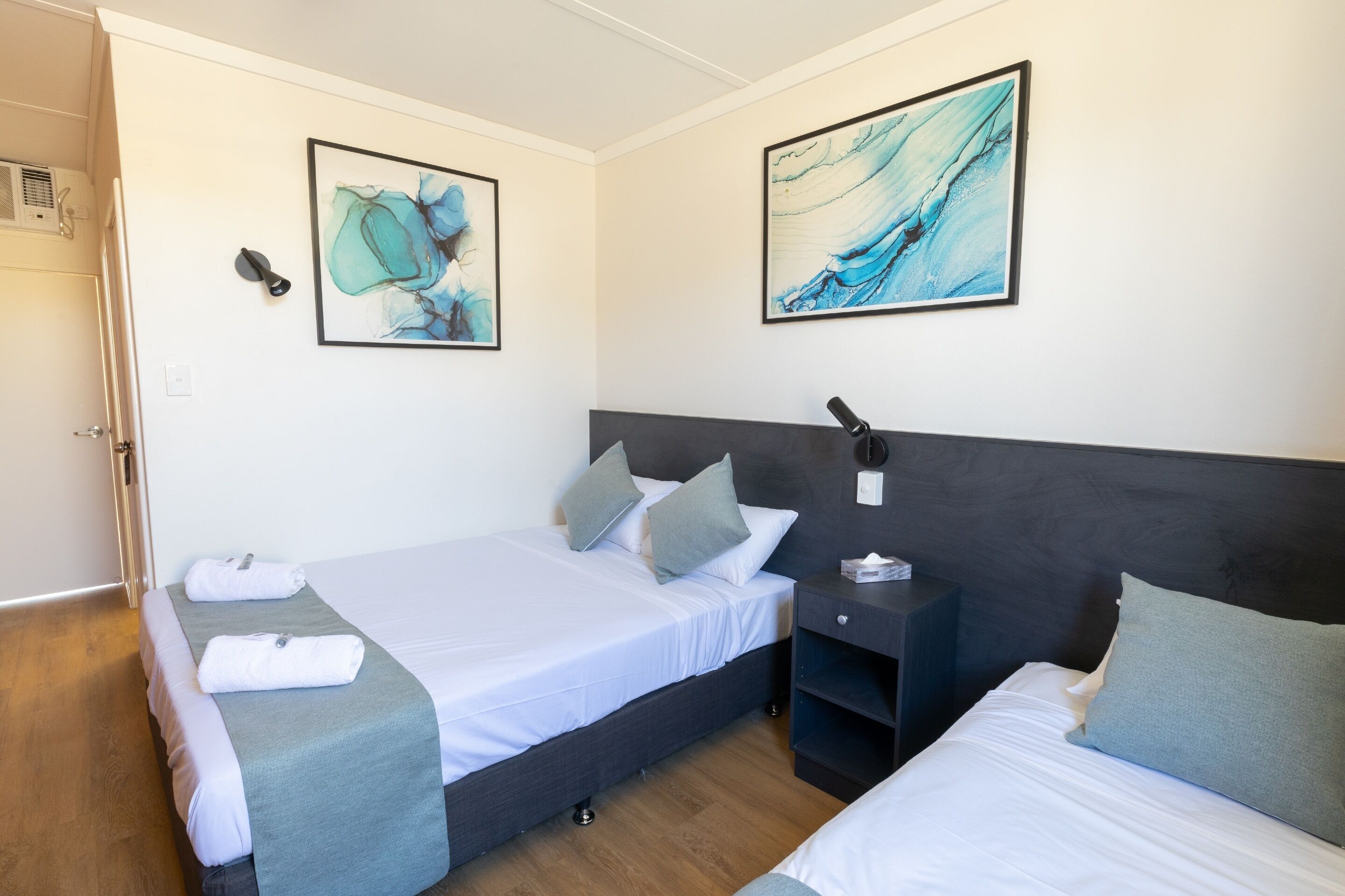 A hotel room with two beds, a dark headboard, a bedside table with a lamp, and artwork on the walls.