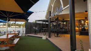 An outdoor seating area with wooden tables and chairs, a white fence, and a building with a sign that reads "The Club". - The Club Hotel (Gladstone)