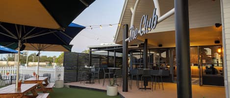Un área de estar al aire libre con mesas y sillas de madera, una cerca blanca y un edificio con un letrero que dice "The Club".