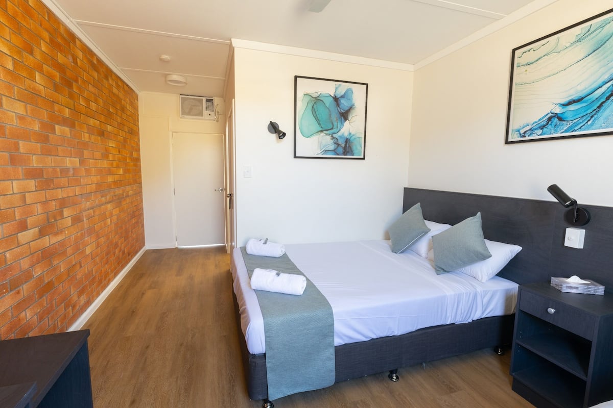 A hotel room with a brick wall, a bed with a grey bedspread, two pillows, a bedside table with a lamp, and two framed artworks on the wall.