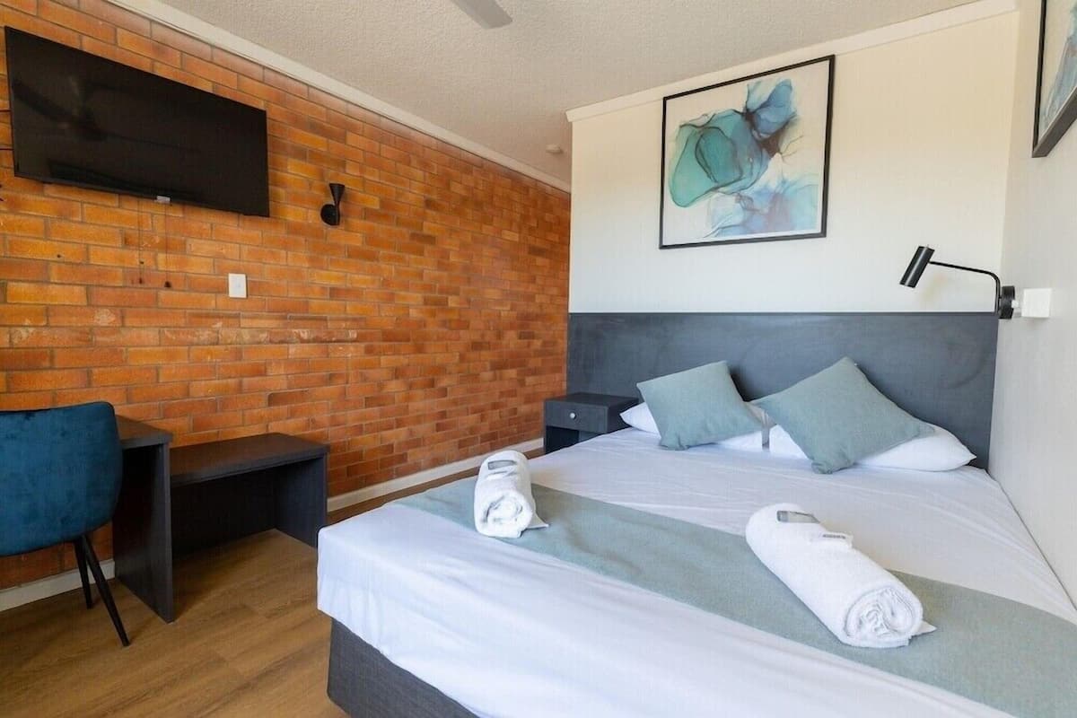 A hotel room with a brick wall, a flat-screen TV, a bed with pillows, a desk, and a chair.