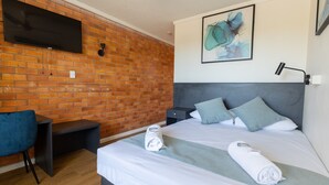 A hotel room with a brick wall, a flat-screen TV mounted above a desk, a bed with a grey headboard, two pillows, and two rolled towels on the bed. - The Club Hotel (Gladstone)