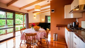 A dining area with a table set for four, wooden chairs, a vase with flowers, and a view of greenery through large windows. - Relax a Lodge (Kerikeri)