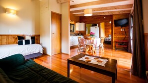 A cozy room with a bed, a green sofa, a wooden dining table with a placemat, and a kitchen area visible through an open door. - Relax a Lodge (Kerikeri)