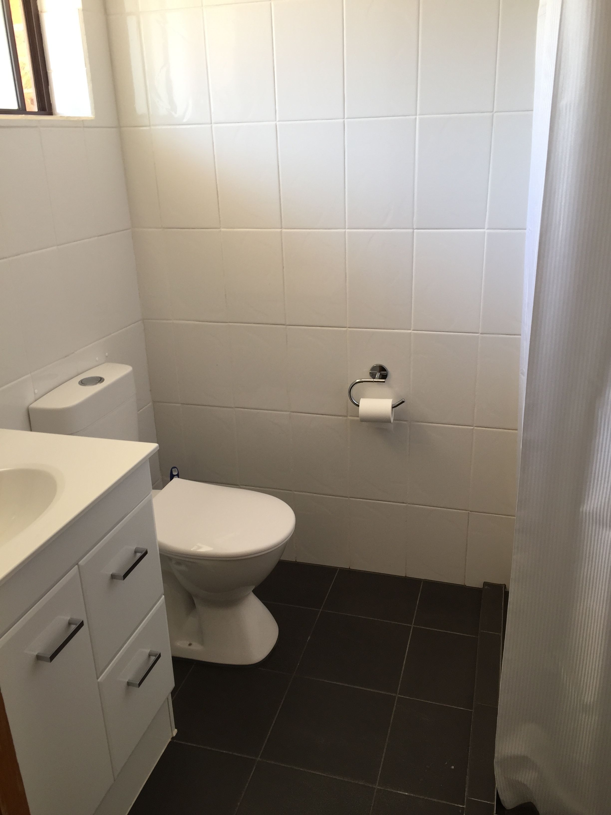 Un baño con inodoro blanco, lavamanos blanco con espejo y suelo de baldosas negras.
