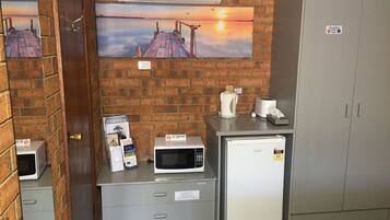 A kitchenette with a microwave, toaster, and coffee maker on a counter, a refrigerator, and a cabinet with drawers. There are two paintings on the wall depicting a sunset over a pier.