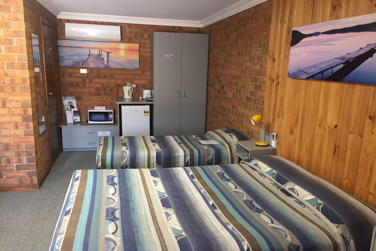 Quarto de hotel compacto com duas camas, uma kitchenette pequena, um guarda-roupa cinzento e uma pintura de um pôr do sol.