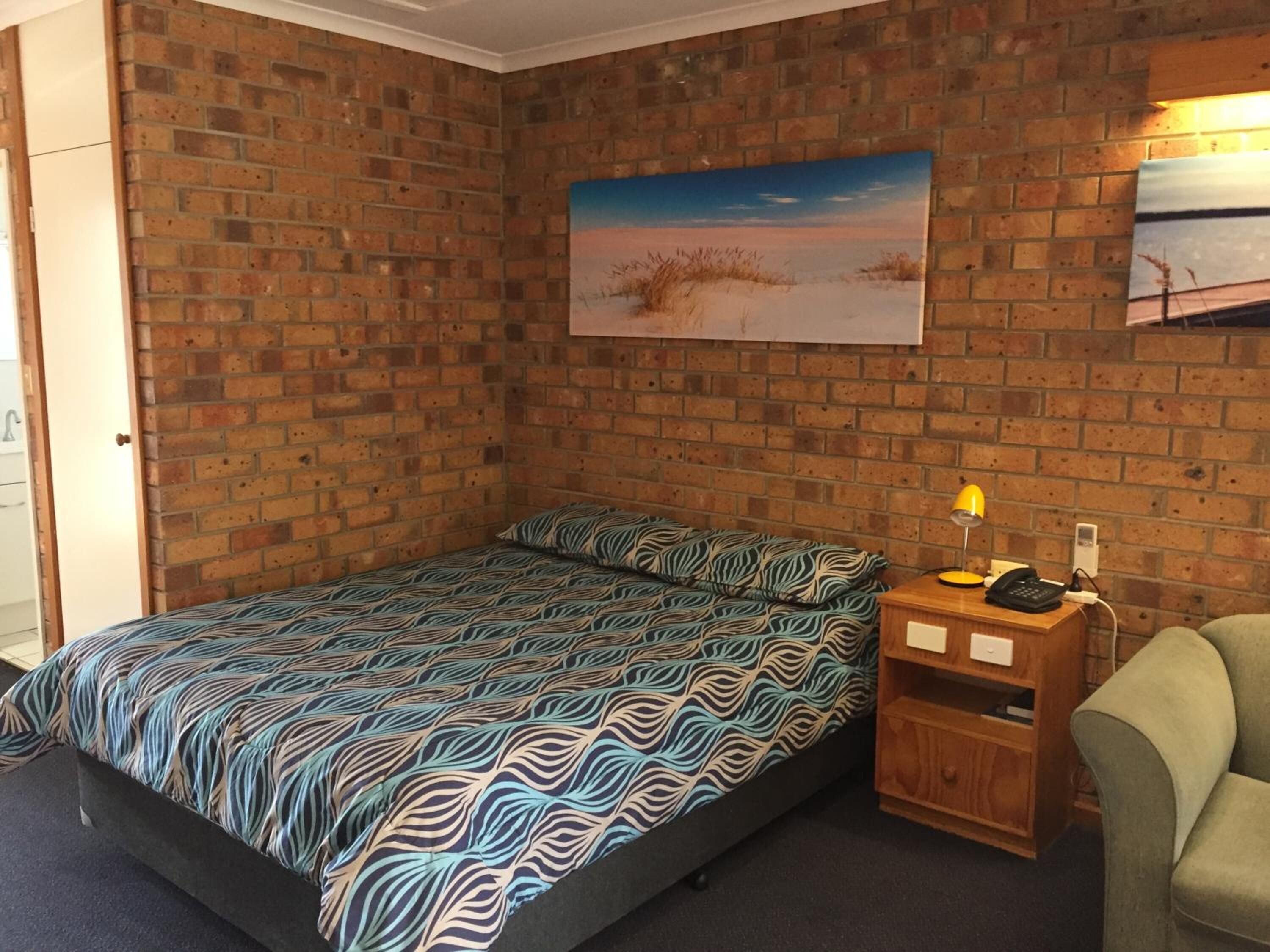 A room with a brick wall, a bed with patterned bedding, a painting of a beach, and a small wooden nightstand with a lamp and phone.