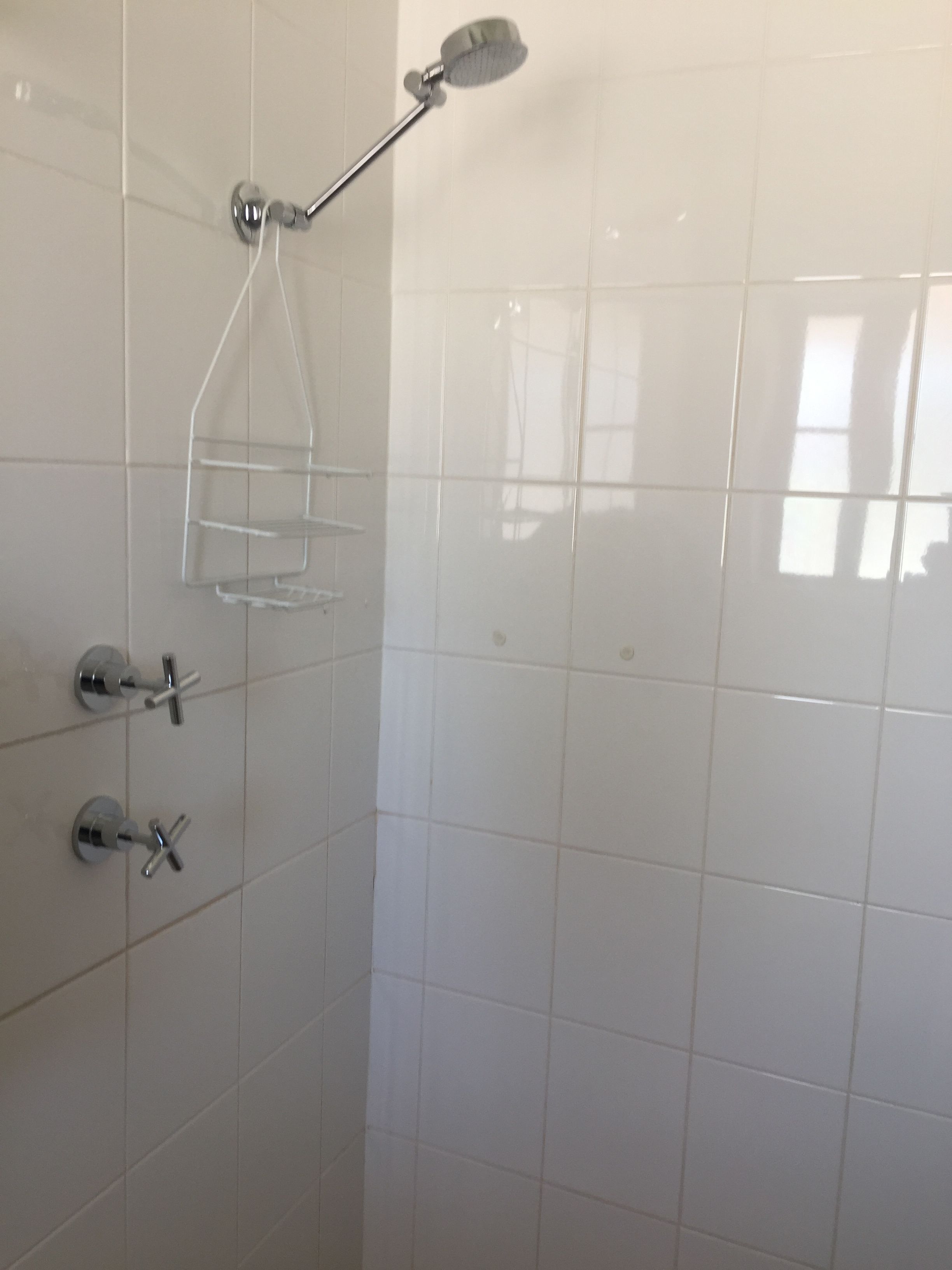 Une salle de bain carrelée de carreaux blancs, une douche avec un pommeau de douche amovible et une étagère en verre.